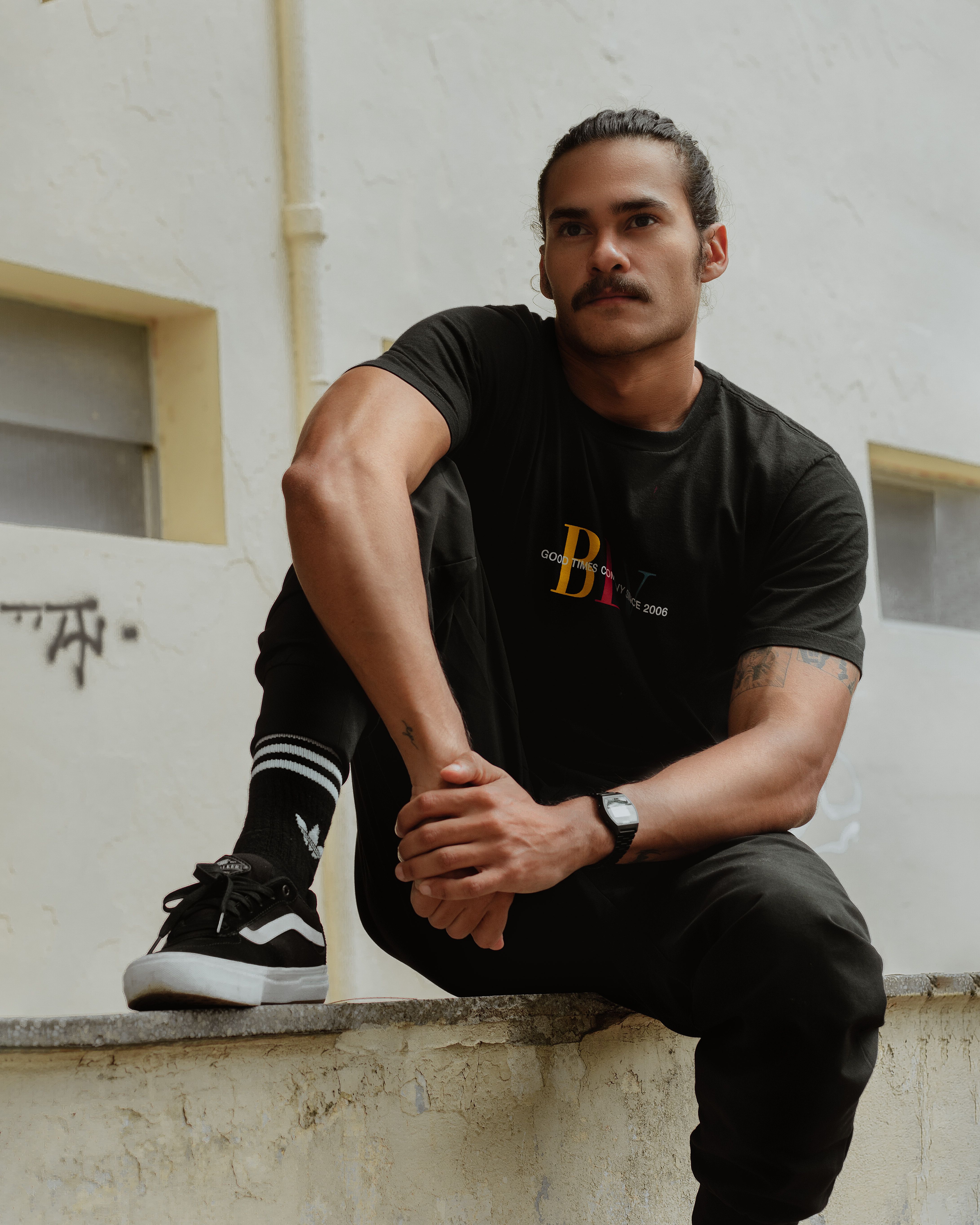 A fashionable young man in streetwear sits on a concrete ledge in an urban setting.