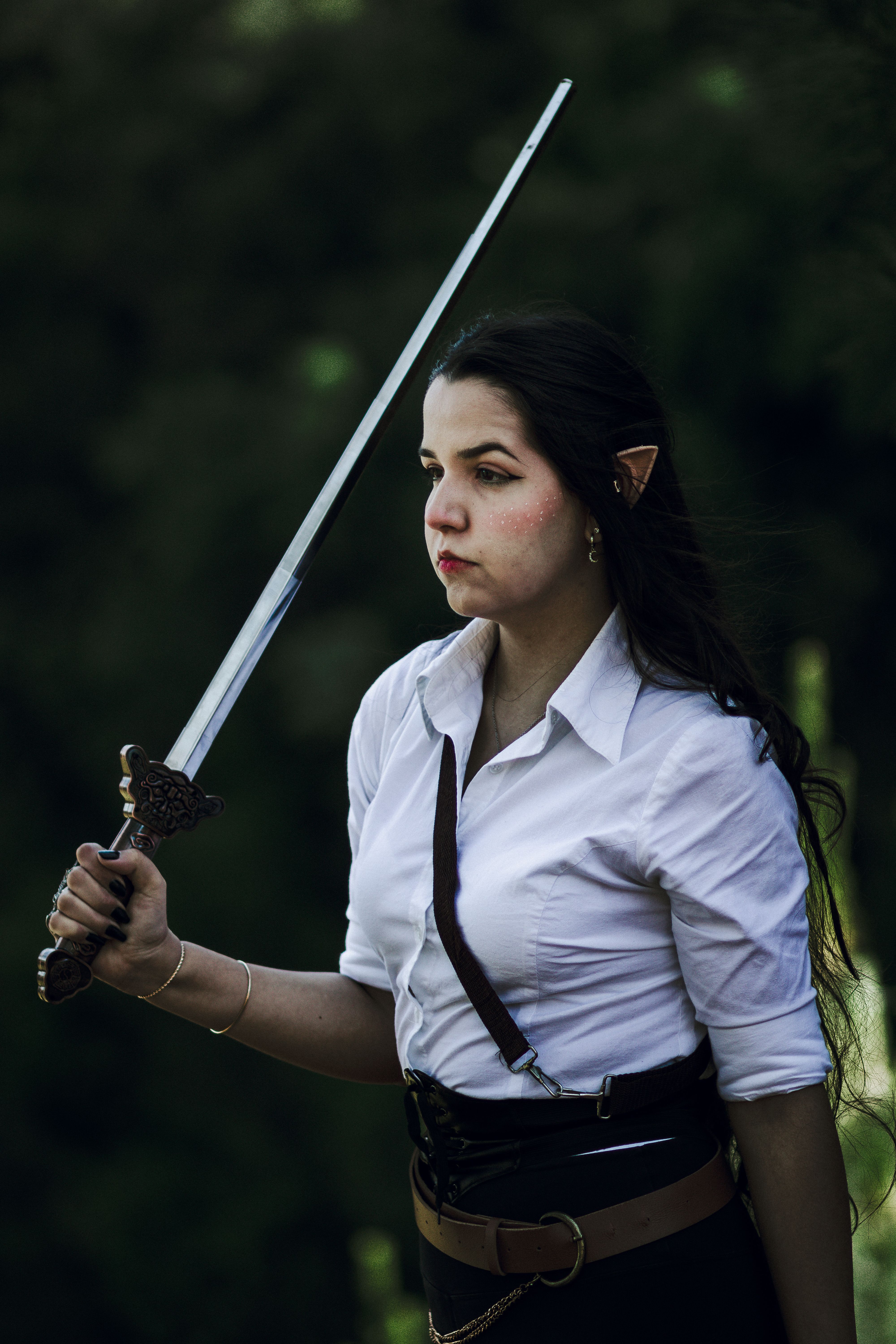 Woman with elf ears in fantasy costume holding a sword in an outdoor setting.