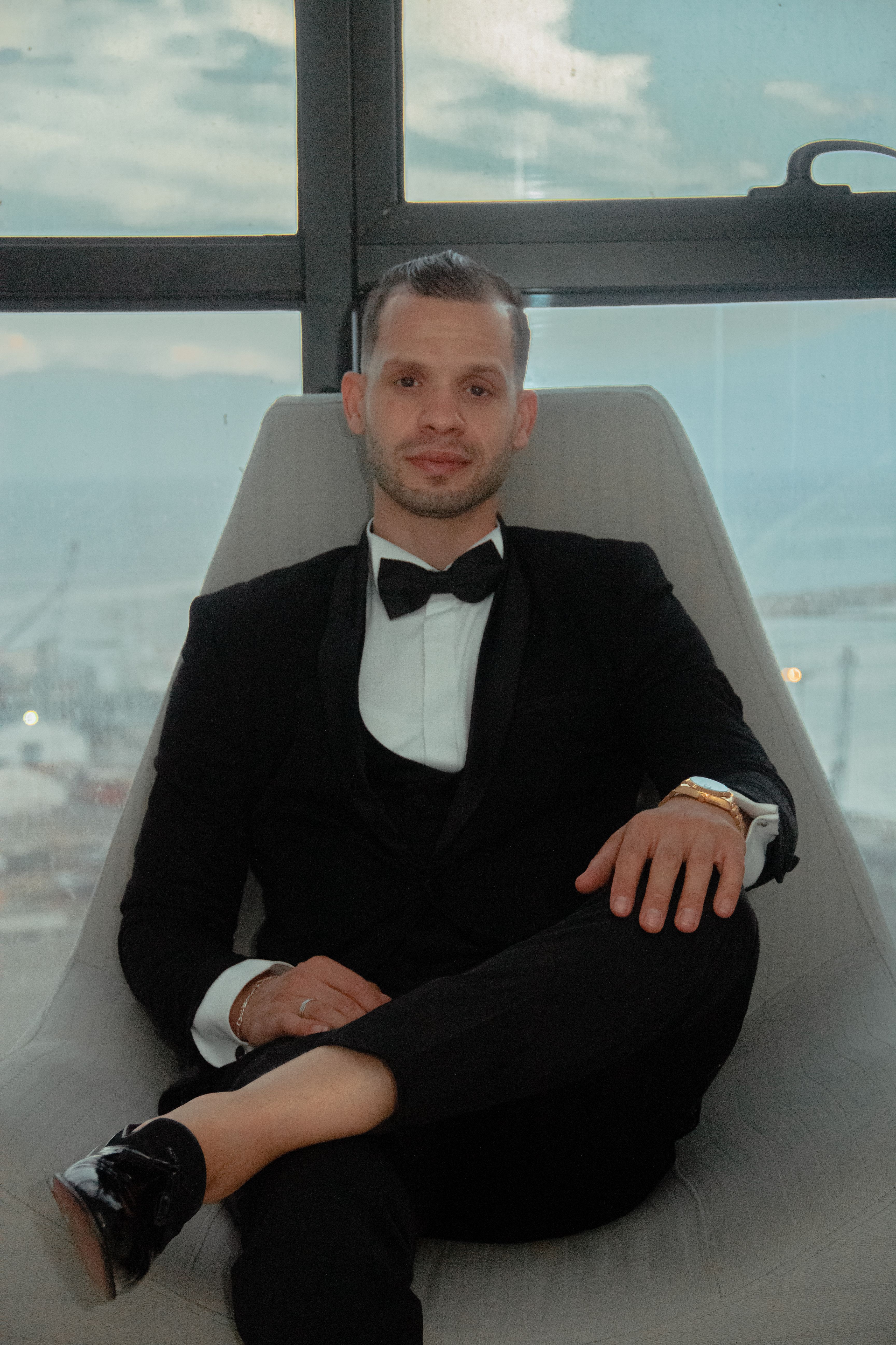 Sophisticated portrait of man in formal attire seated by a large window with an industrial view.