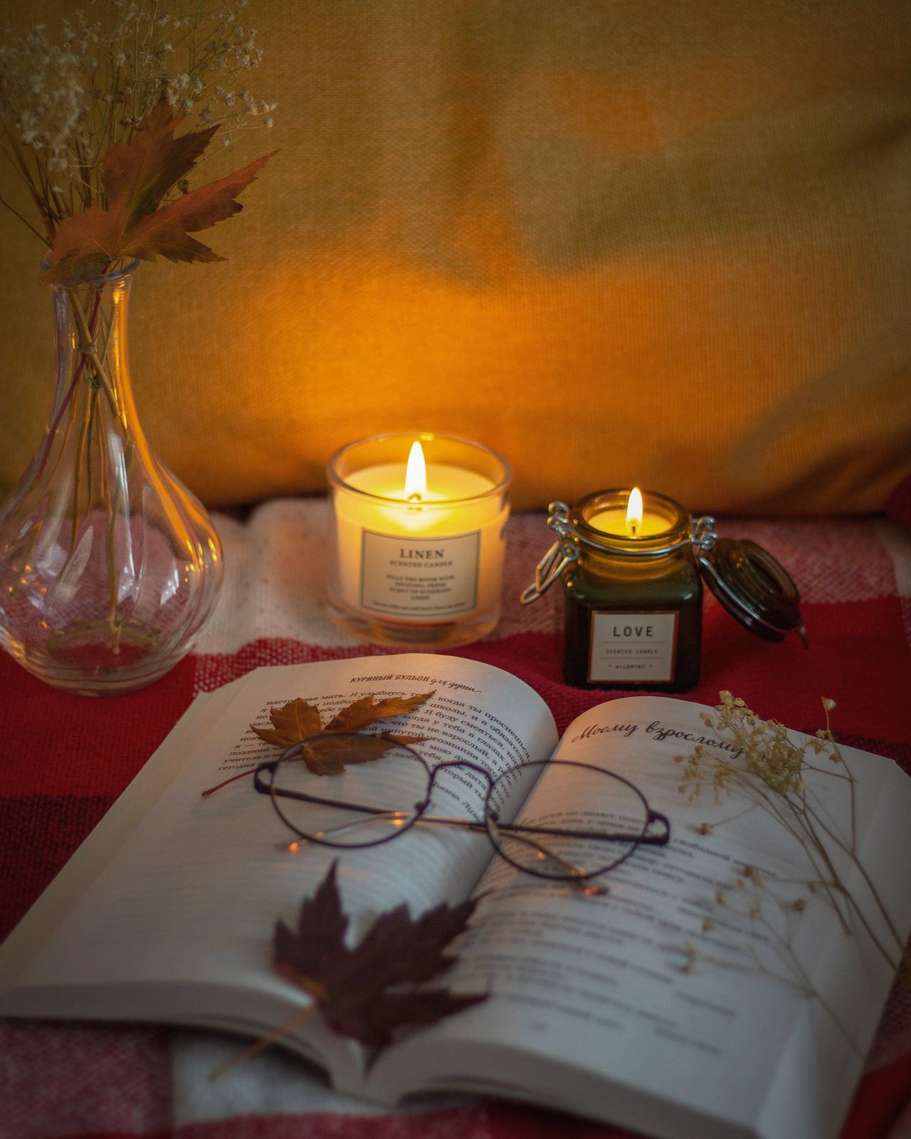 Lit candles, an open book, reading glasses, and autumn leaves on a cozy surface