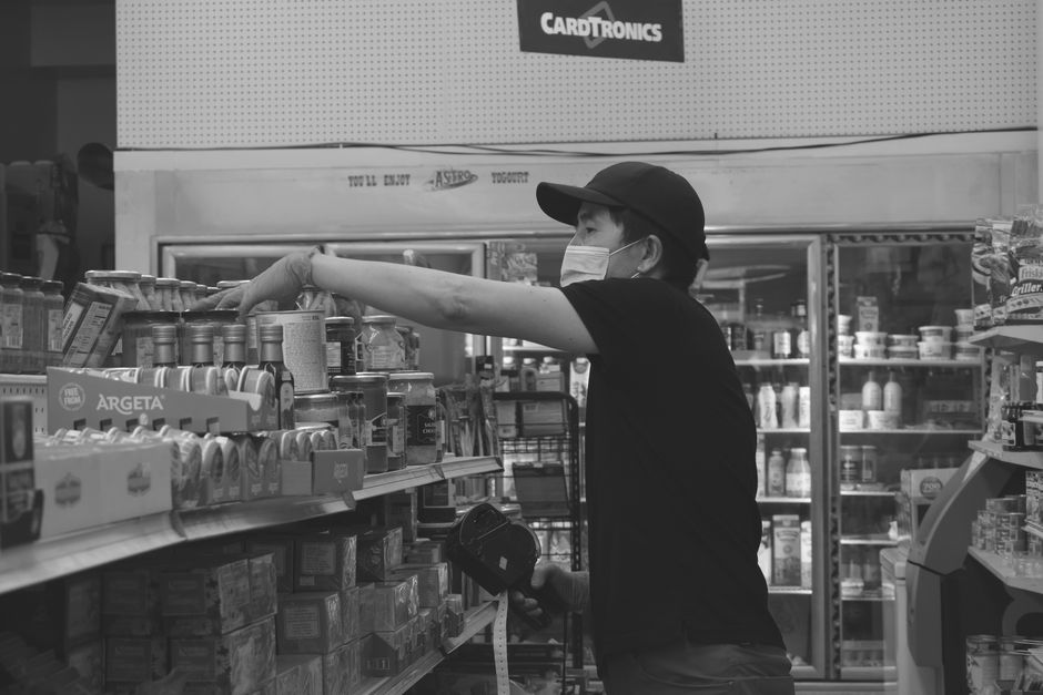 Monochrome photo of a masked worker organizing canned goods in a grocery store.