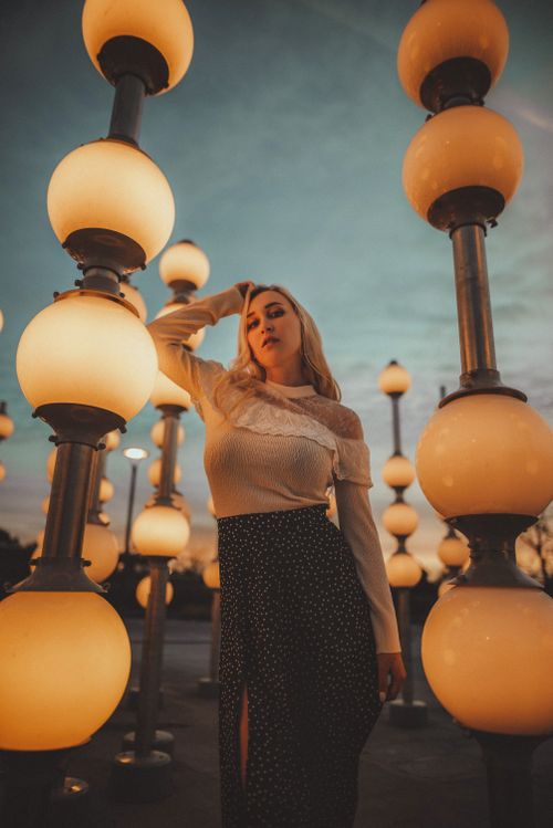 Free Stylish woman poses among glowing orb lamps at sunset, showcasing fashion and elegance. Stock Photo