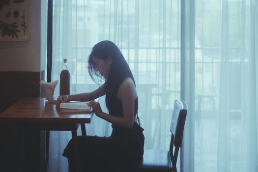 A woman engrossed in a book at a cafe table with gentle lighting through curtains, creating a serene atmosphere.