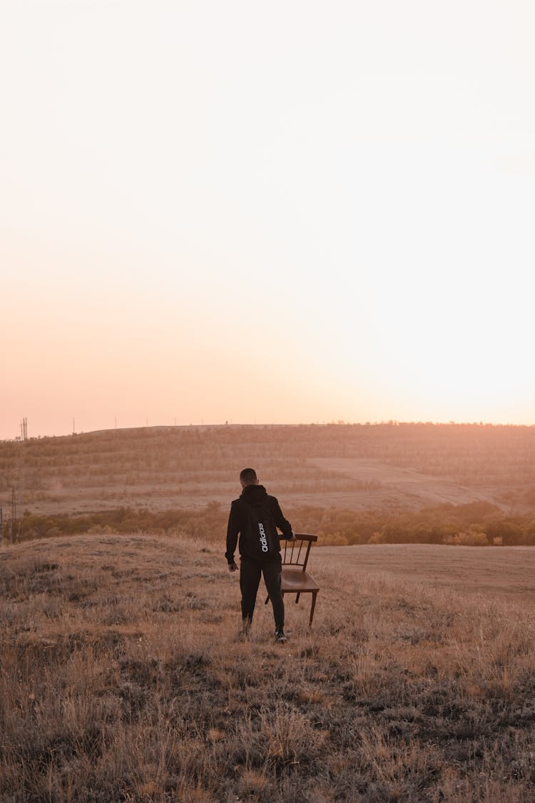 Man Carrying A Wooden Chair On Grass Field During Sunset