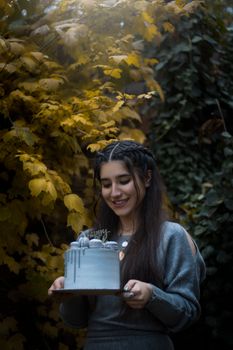 Smiling young woman holds a birthday cake in a lush outdoor setting with autumn leaves.