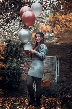 Smiling woman holding balloons and cake in a rustic autumn setting, perfect for celebrating birthdays.