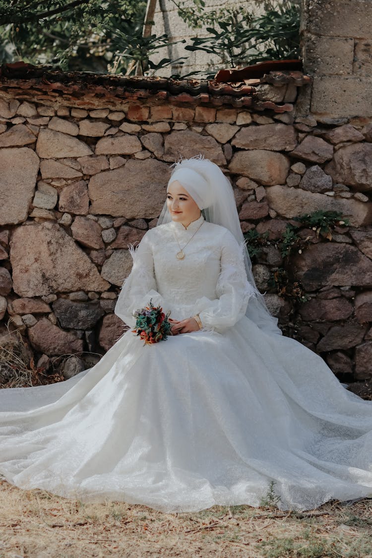 Smiling Bride In Wedding Dress And Veil Sitting By Wall In Garden And Smiling