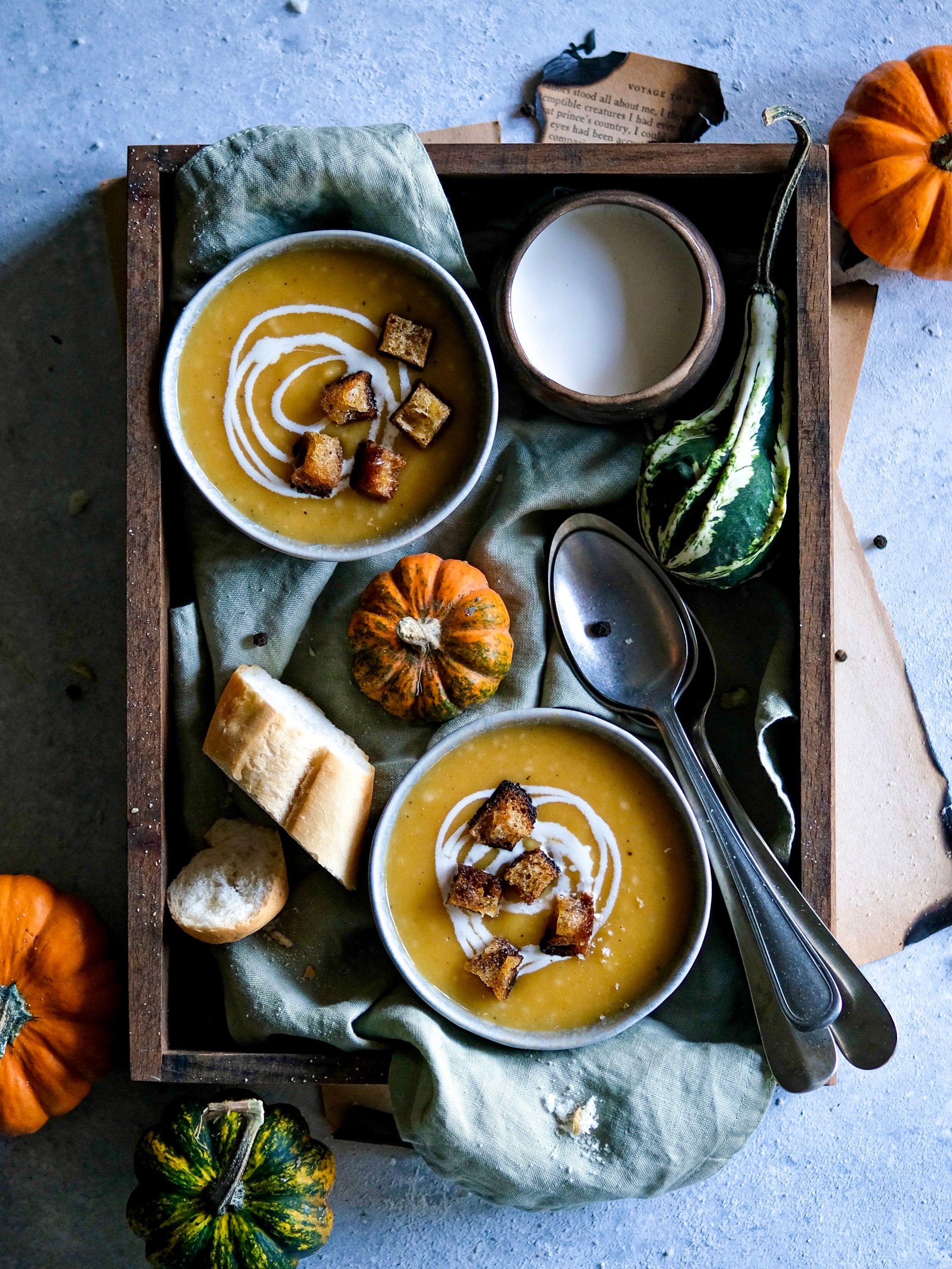Warm and inviting fall-themed pumpkin soup served with croutons, showcased with gourds and bread in a wooden tray.