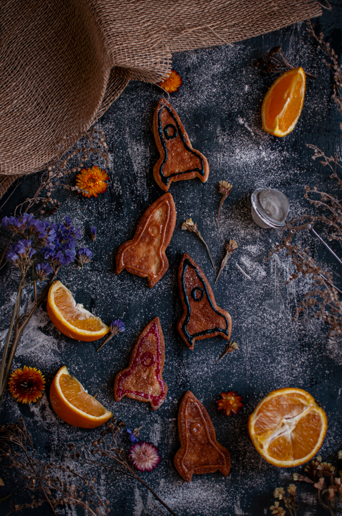 Free Artistic flat lay featuring rocket-shaped snacks with colorful flowers and sliced oranges on a textured surface. Stock Photo