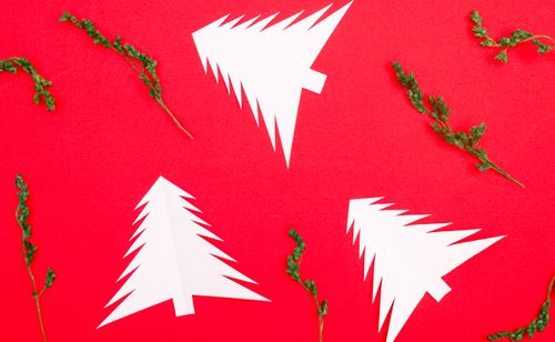 Free White paper Christmas tree cutouts arranged on a vibrant red background with green floral elements. Stock Photo
