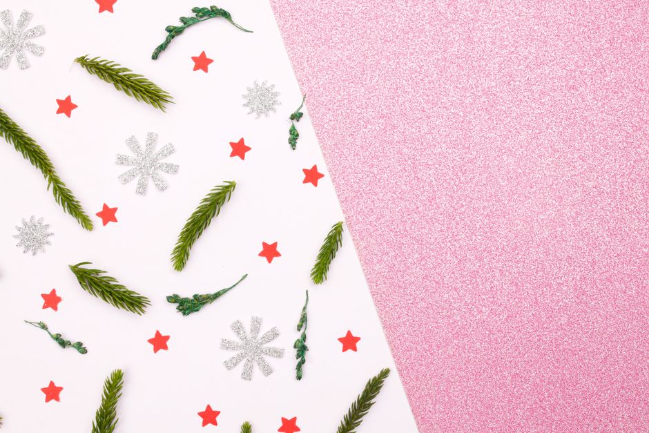 Christmas flat lay with green sprigs, red stars, and silver sparkles on pink and white paper.