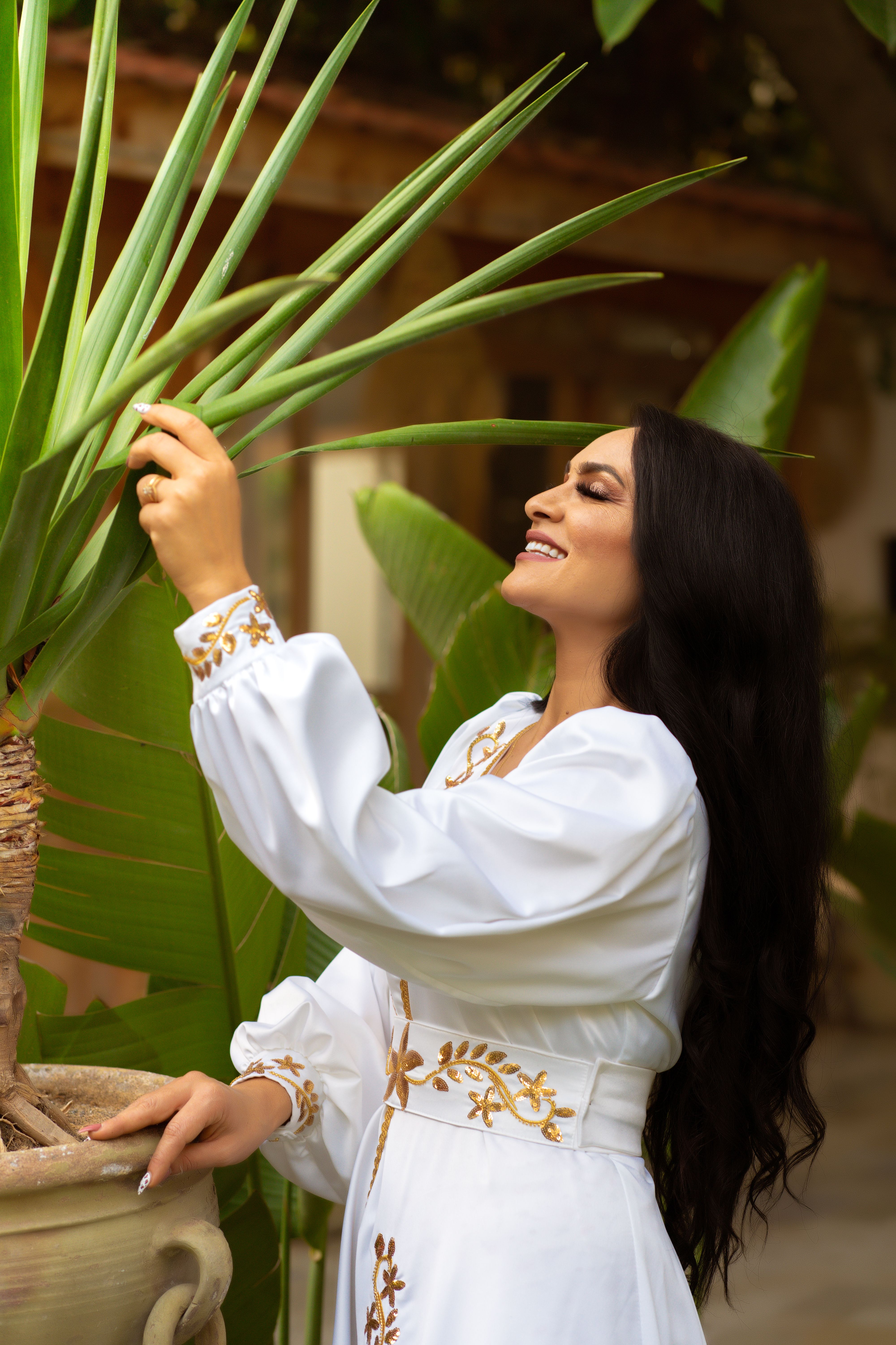 Smiling woman in a white embroidered dress interacts with tropical plant outdoors.