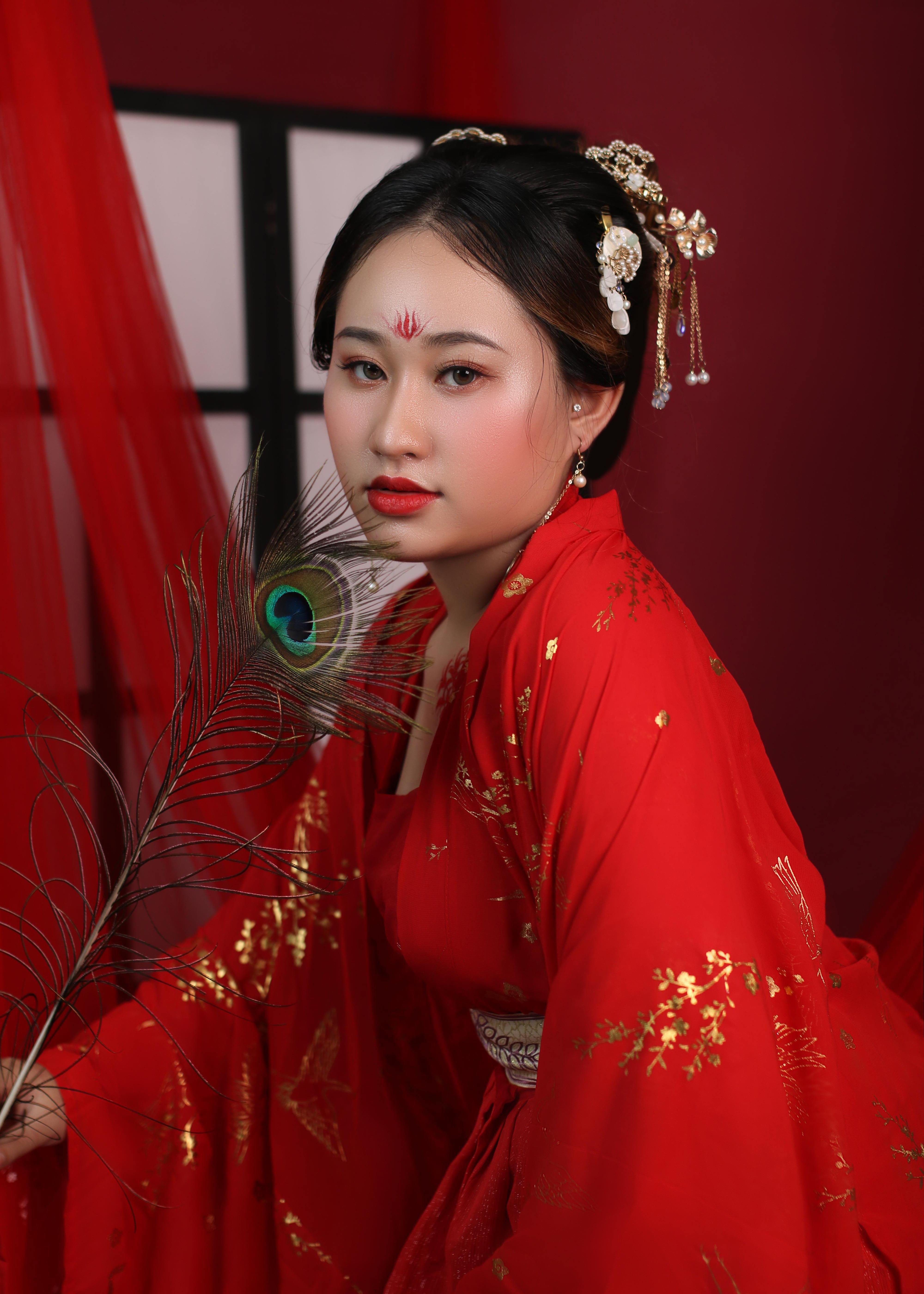 Portrait of a woman in traditional red attire holding a peacock feather, exuding elegance.