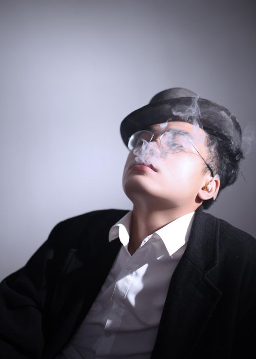 Free Asian man wearing a black hat and suit exhales smoke in an indoor studio setting. Stock Photo