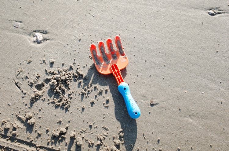 A Blue And Orange Plastic Rake On Gray Sand