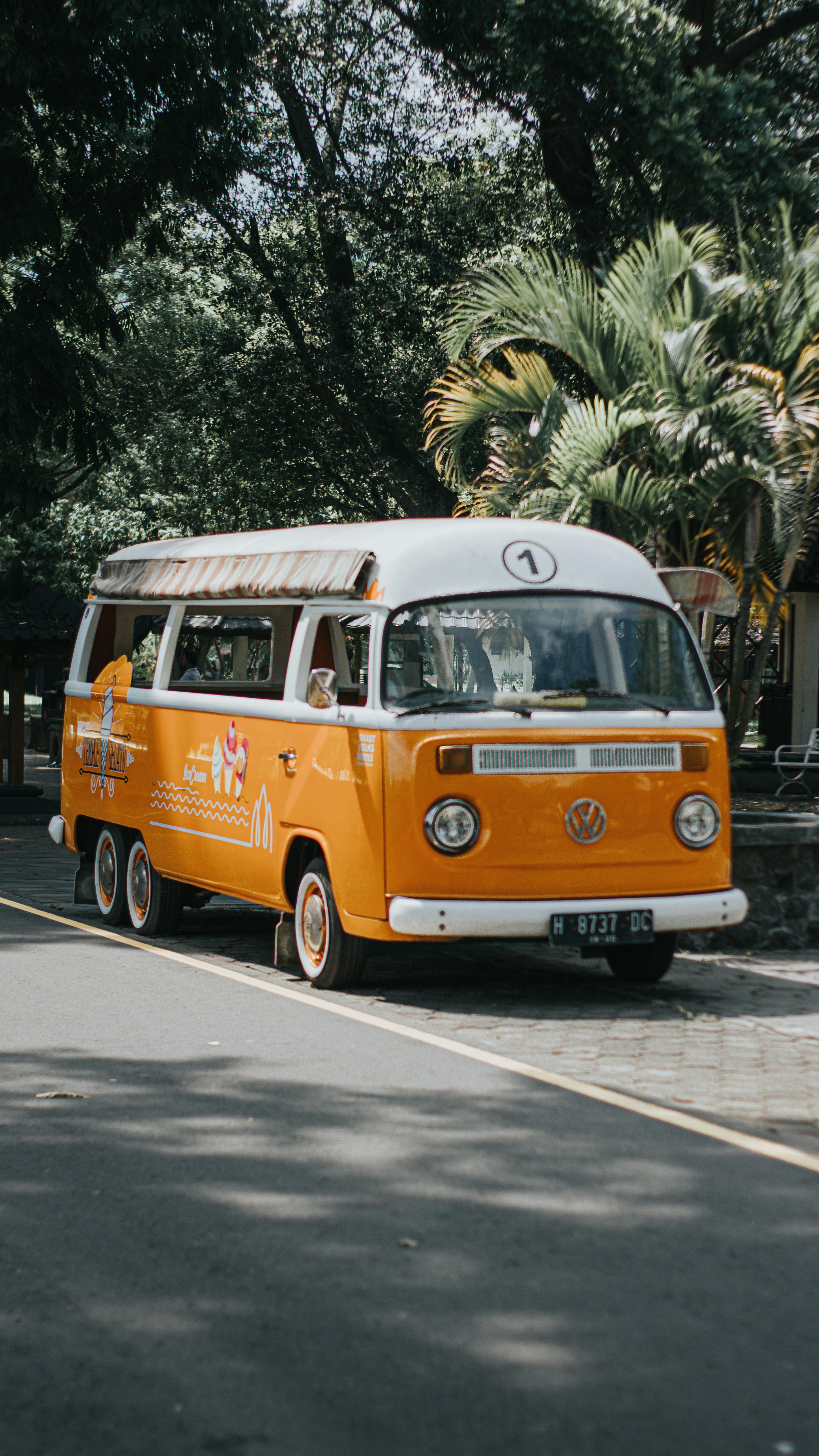 Orange vintage bus parked on a tree-lined road with summer trees in the background.