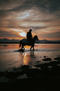A lone cowboy silhouettes against a dramatic sunset while riding along a serene lake.