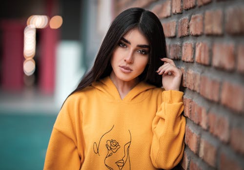 Free Shallow Focus of a Beautiful Woman in Yellow Hoodie Sweater Leaning on Brick Wall Stock Photo