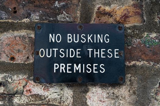 Black sign stating 'No Busking' affixed to a textured brick wall surface.