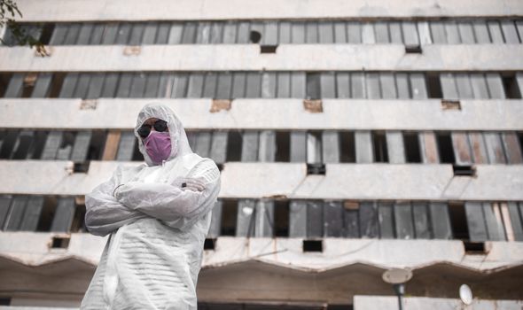 Person wearing PPE, mask, and sunglasses standing in front of a derelict building.