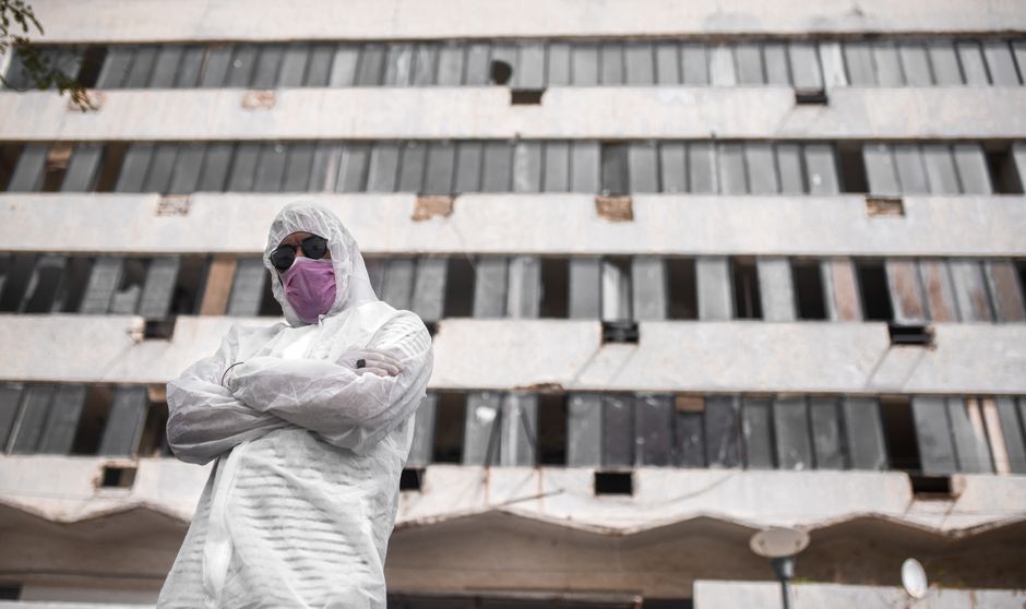 Person wearing PPE, mask, and sunglasses standing in front of a derelict building.