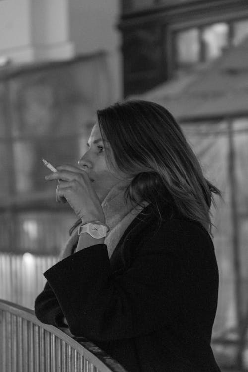 Grayscale Photo of a Woman in Black Coat Smoking a Cigarette