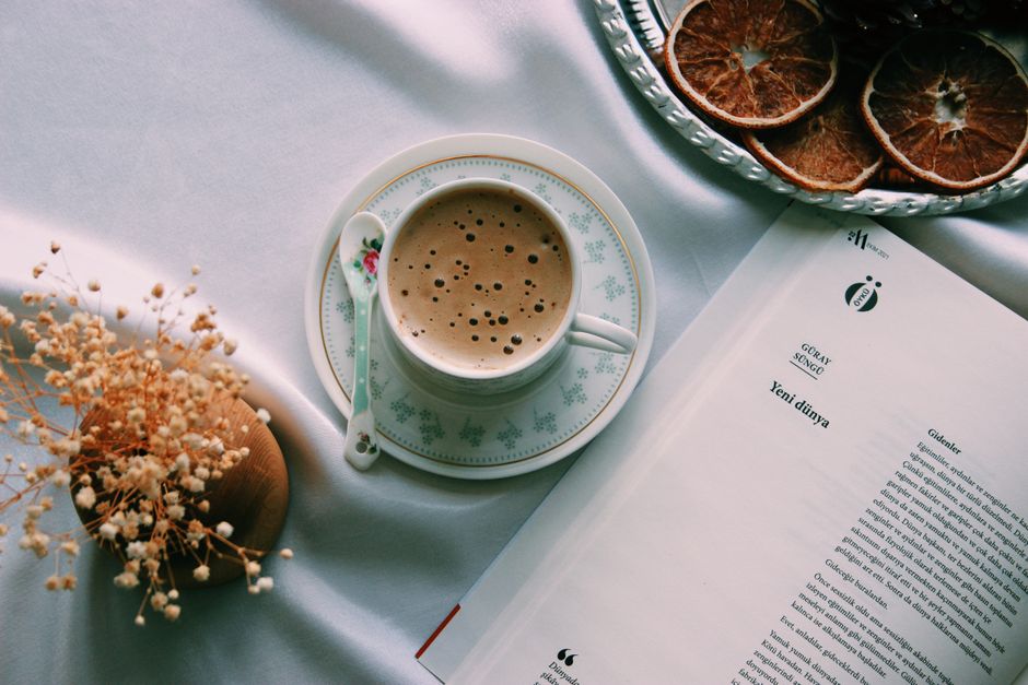 A flat lay of coffee with dried orange slices, flowers, and an open book on a white tablecloth.