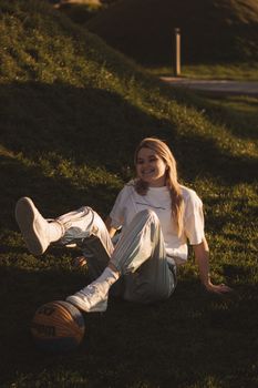 Smiling young woman sitting on grass with a basketball, enjoying a sunny day outdoors.