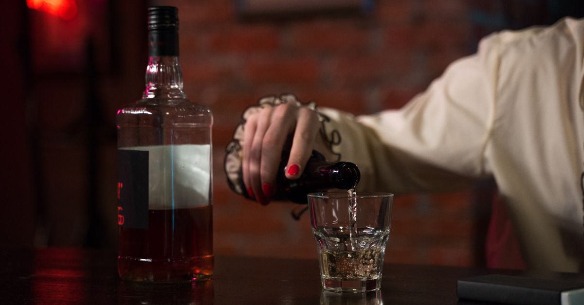 A hand with red nail polish pouring whiskey into a glass at a bar with a brick wall backdrop.
