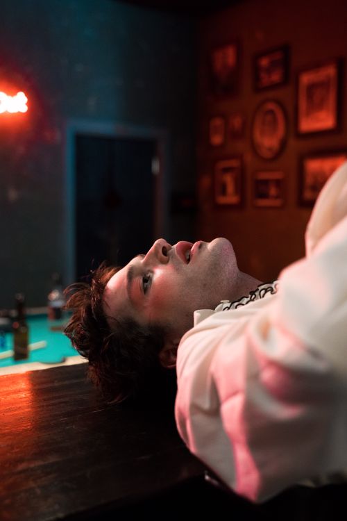 Free A man lying on a bar counter, surrounded by moody light and framed wall art. Stock Photo