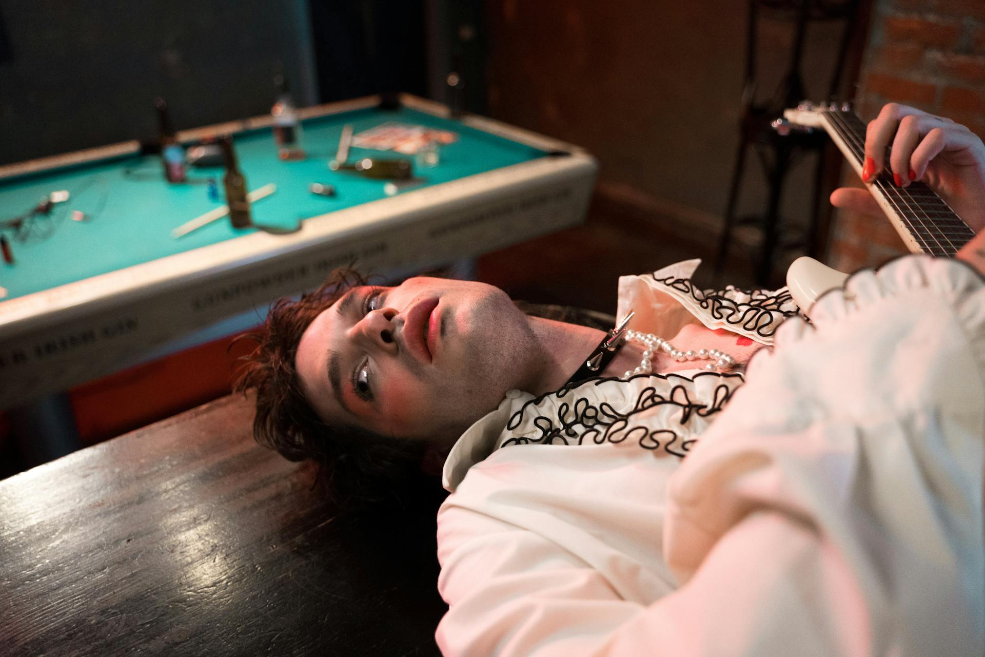 A laid-back rock musician in a stylish outfit, lying near a pool table indoors.