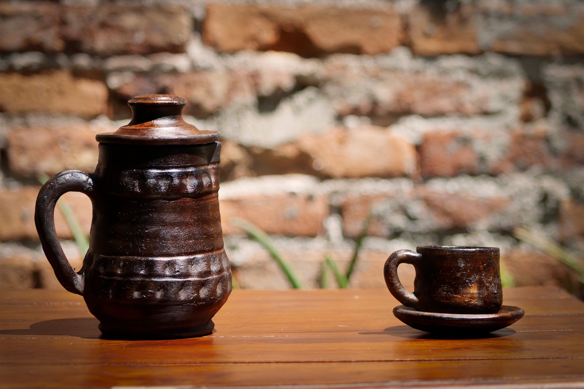 A rustic ceramic teapot and cup set on a wooden table against a brick wall, basking in sunlight.