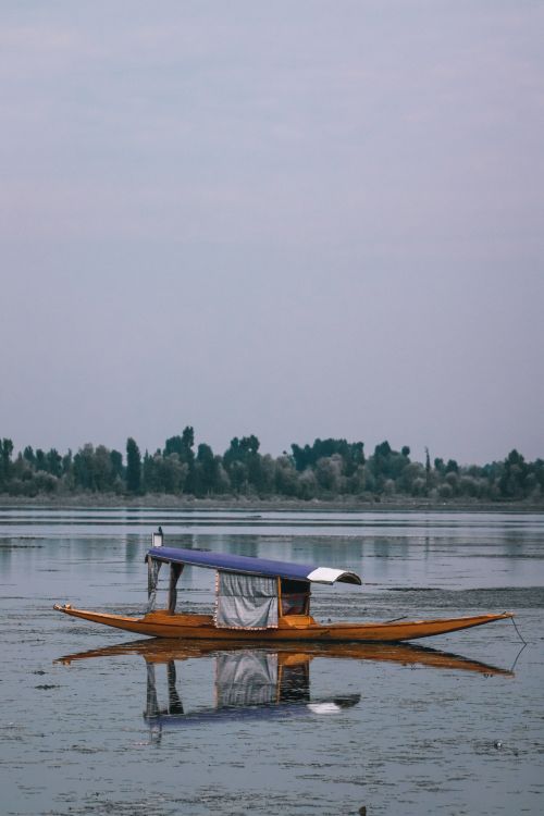 Free A traditional wooden boat reflects in calm lake waters, surrounded by trees under a soft, overcast sky. Stock Photo