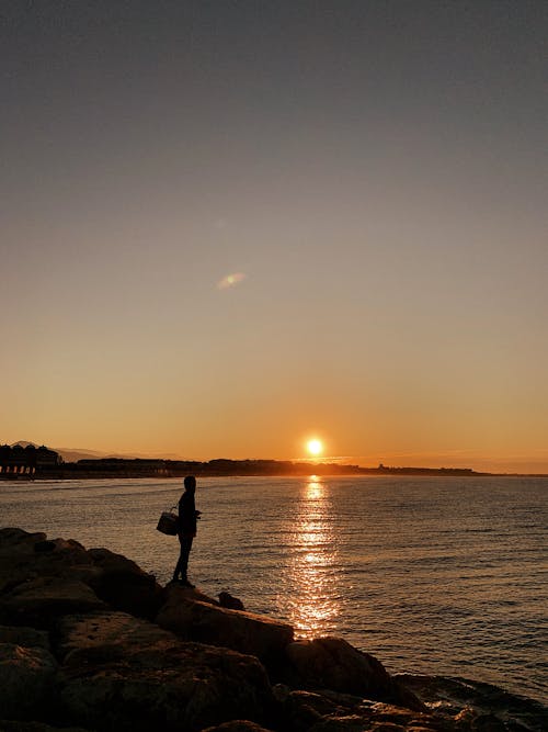 Silhouette of a Person Standing on a Rock Near Body of Water