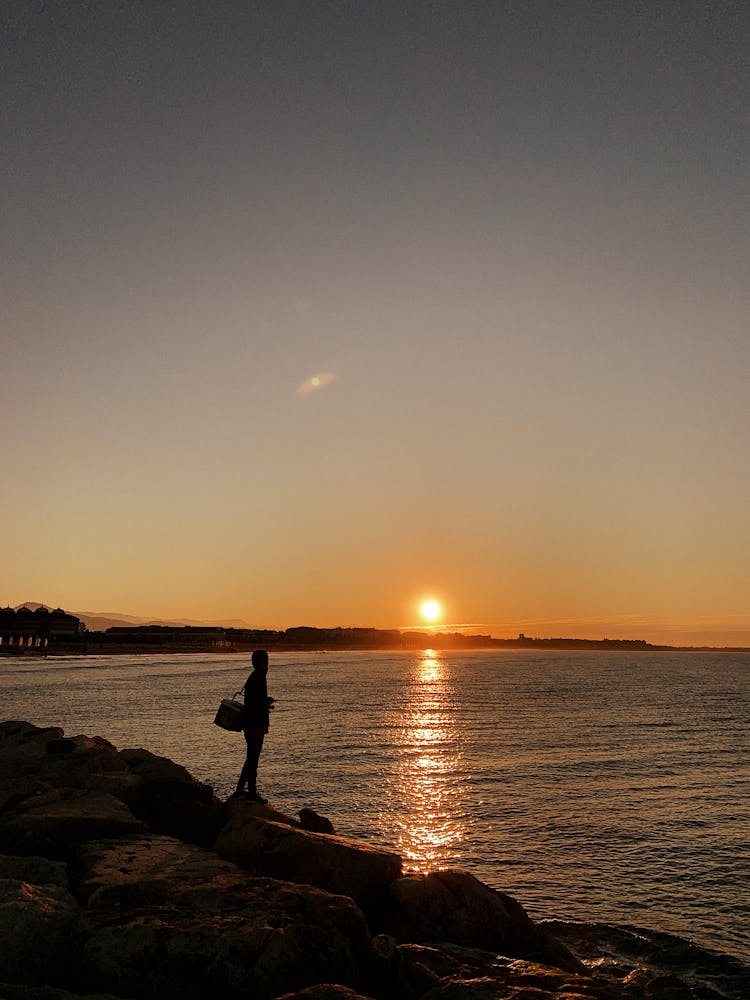 Silhouette Of A Person Standing On A Rock Near Body Of Water