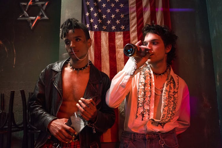 Young Men Holding Liquor Bottles At A Party And Standing On The Background Of The US Flag 