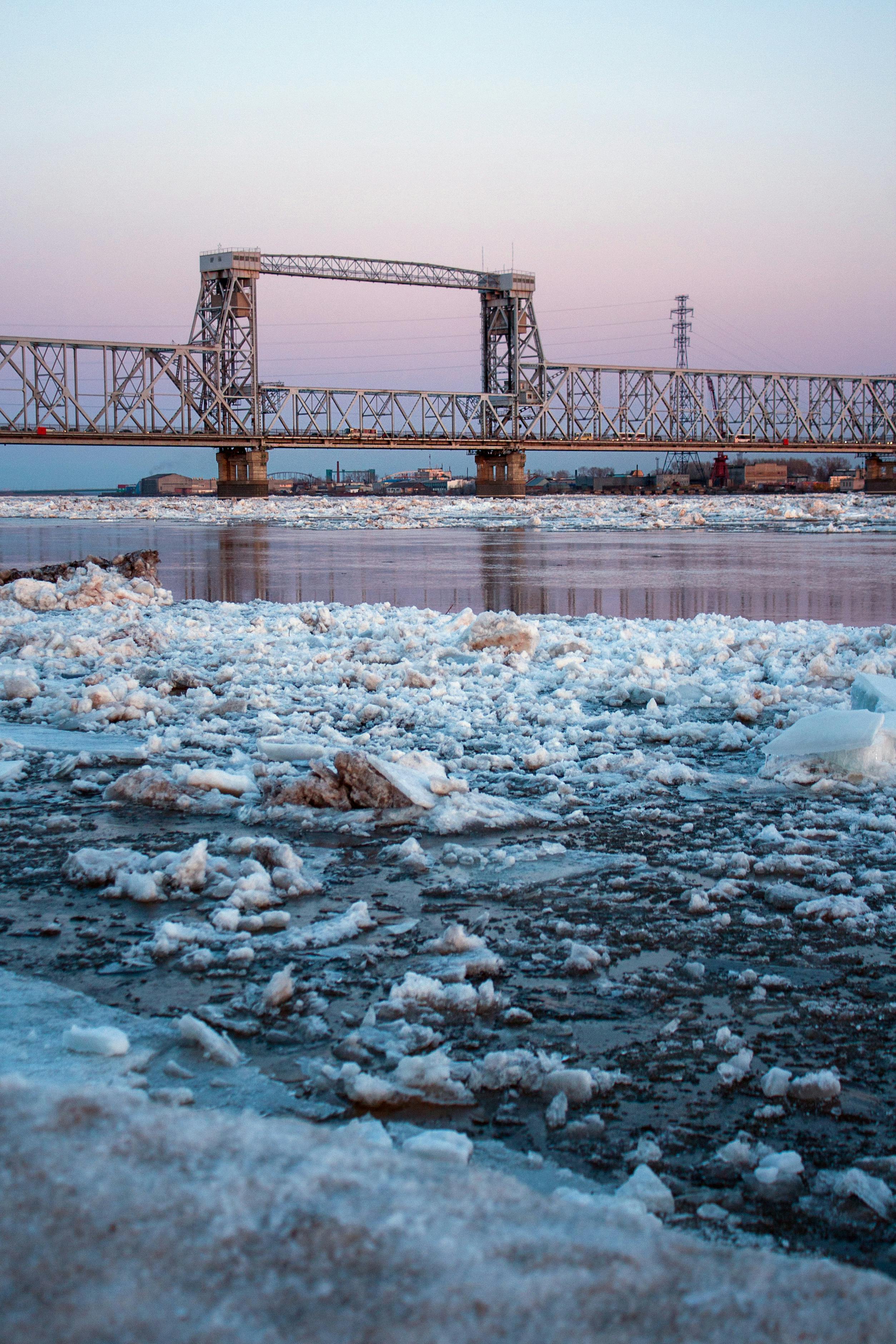 A steel bridge spans a frozen river covered with ice chunks during winter twilight in Arkhangelsk, Russia.
