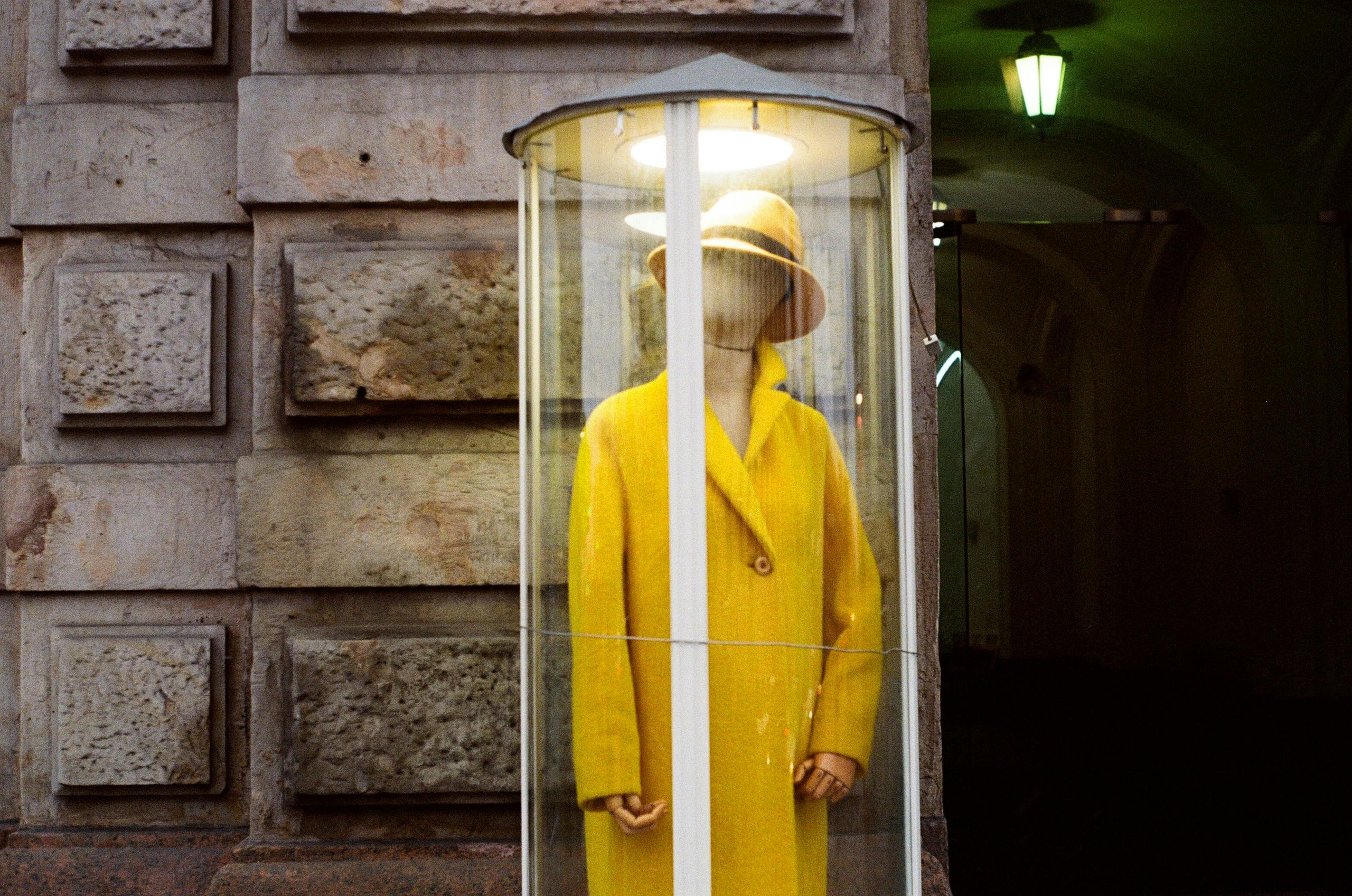 A mannequin in a bright yellow coat encased in glass against a stone wall.