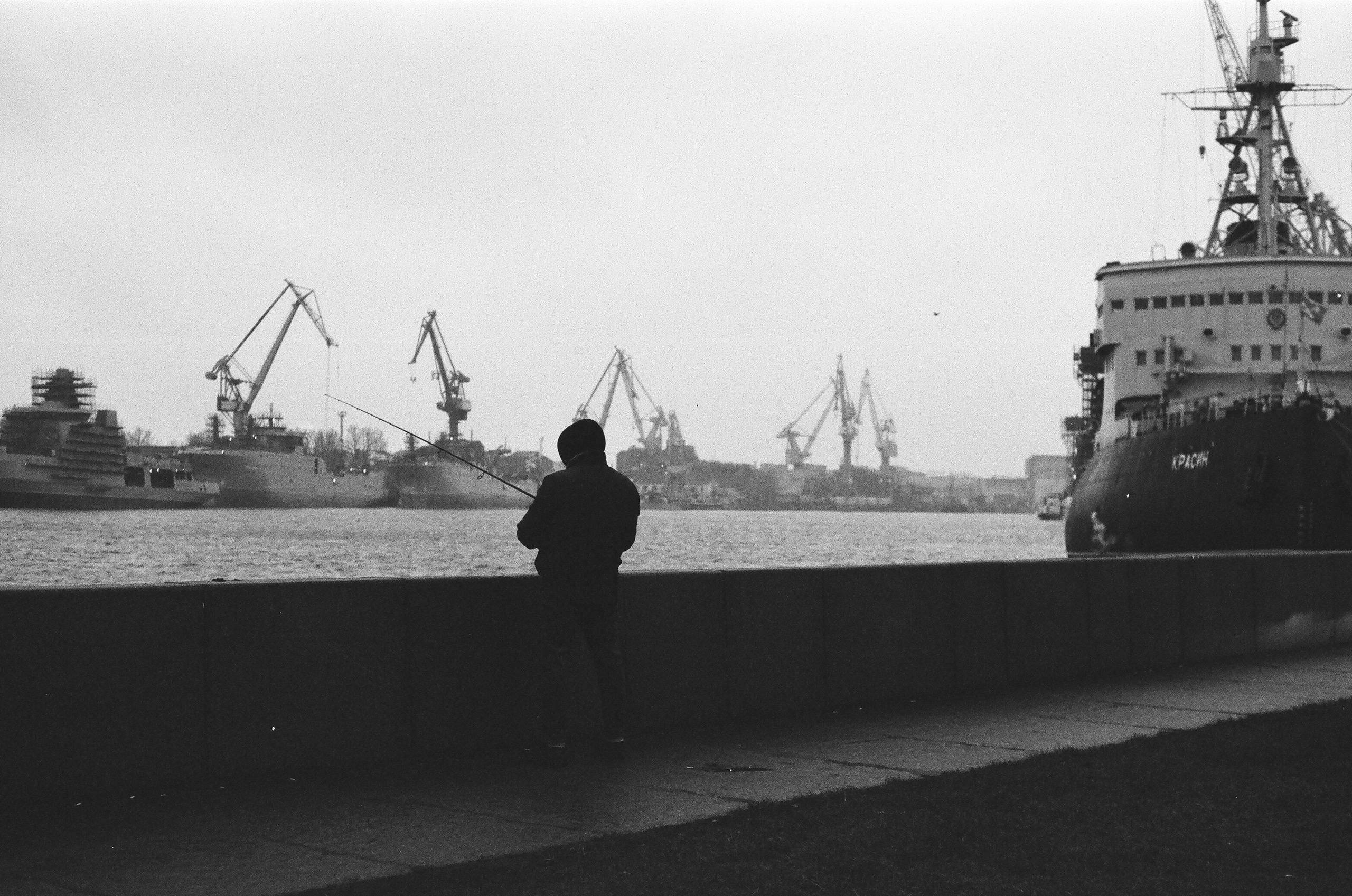 A person fishes by the harbor in Saint Petersburg, capturing a serene moment in monochrome.
