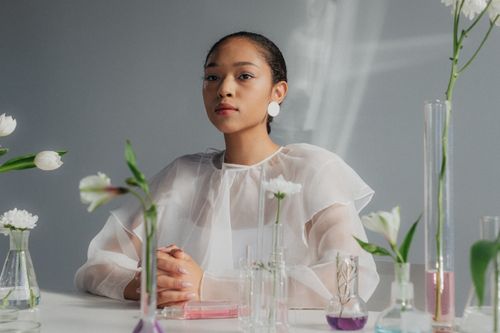 Free A woman in a sheer blouse sits in a botanical lab with flowers and glassware. Stock Photo