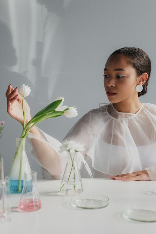 Free A woman in a diaphanous top interacts with tulips in scientific glassware, merging art and science. Stock Photo