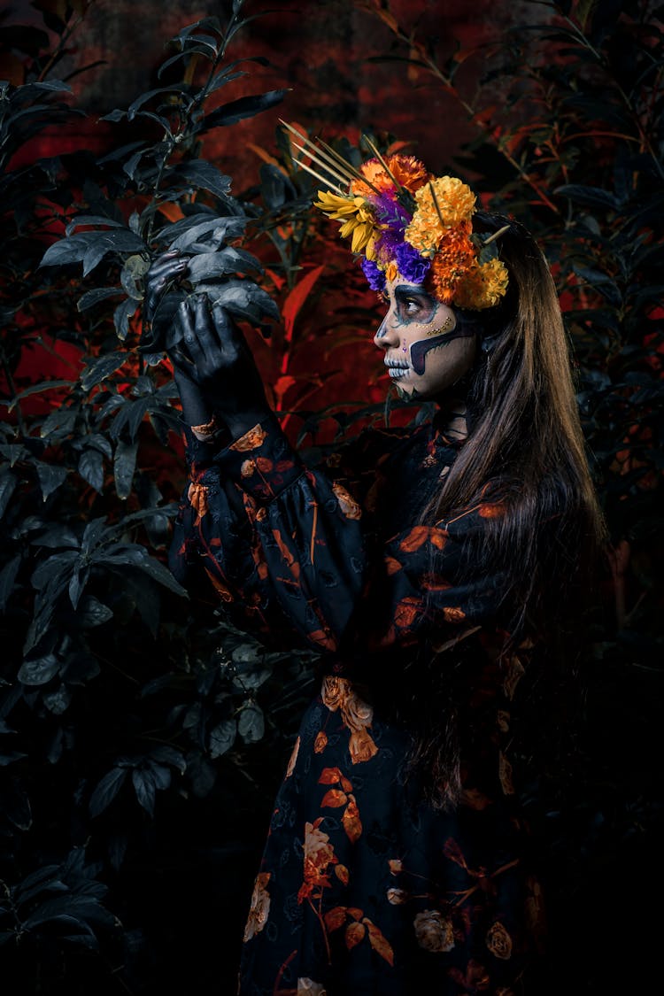 A Woman With Painted Face Wearing A Floral Headdress Looking At The Leaves Of A Plant