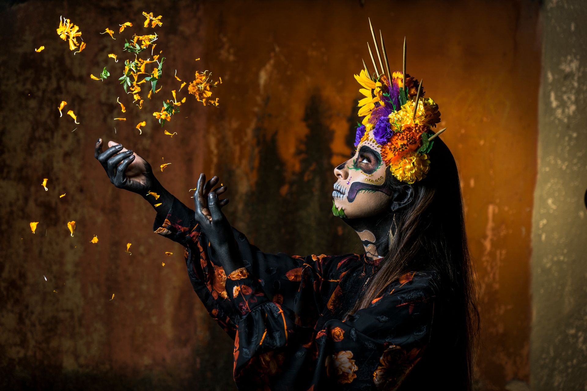 A woman in Day of the Dead makeup and floral crown celebrates with colorful flower petals.