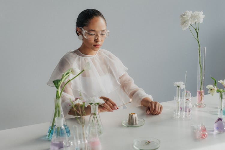 Female Scientist In Goggles Standing At Table With Lit Match In Hand