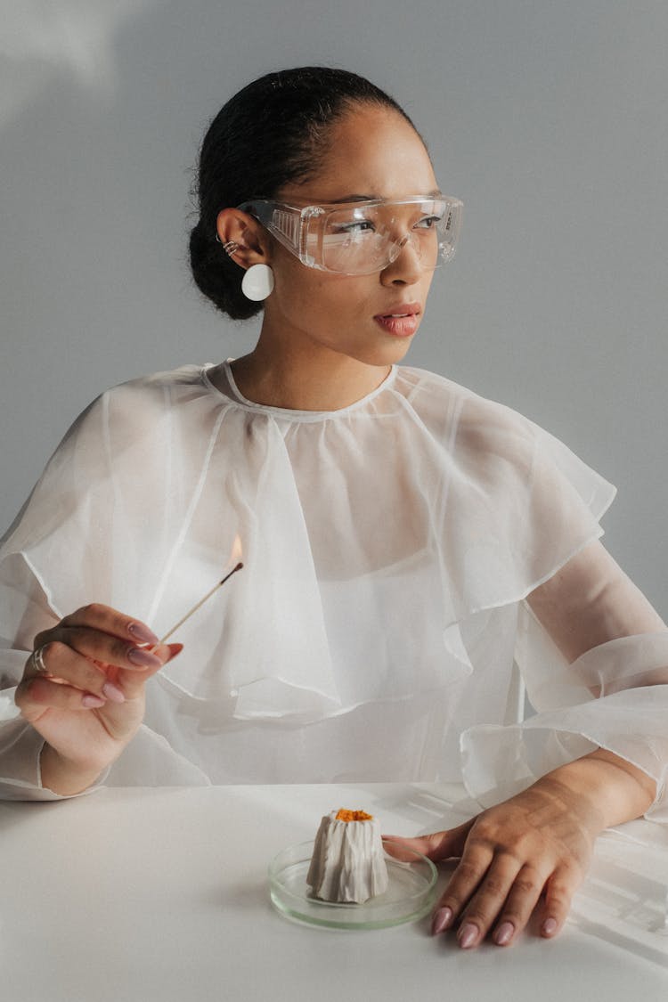 Woman Pretending Scientist Sitting At White Table And Holding Lit Match In Hand