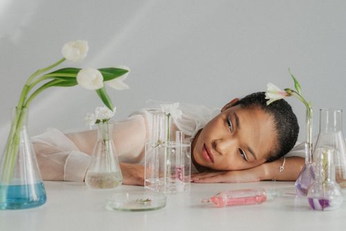 Free A woman thoughtfully rests among flowers and color flasks in a serene laboratory setting. Stock Photo