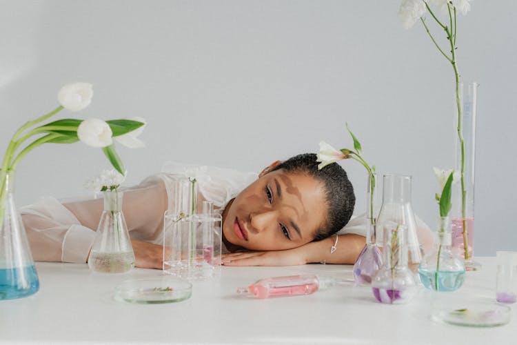 Woman Falling Asleep With Head On Table Among Flowers In Laboratory Flasks