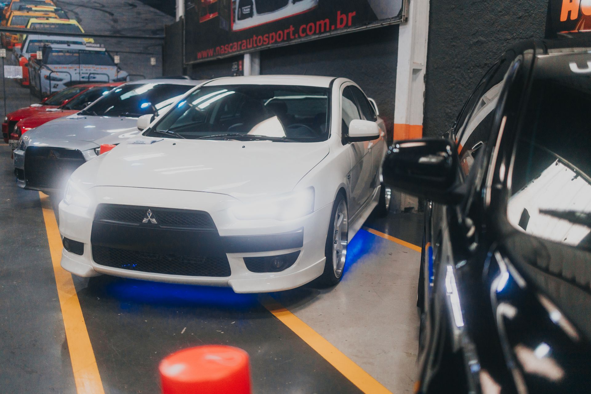 Parked Mitsubishi Lancer Evolution with illuminated headlights inside a garage showcasing sports cars.