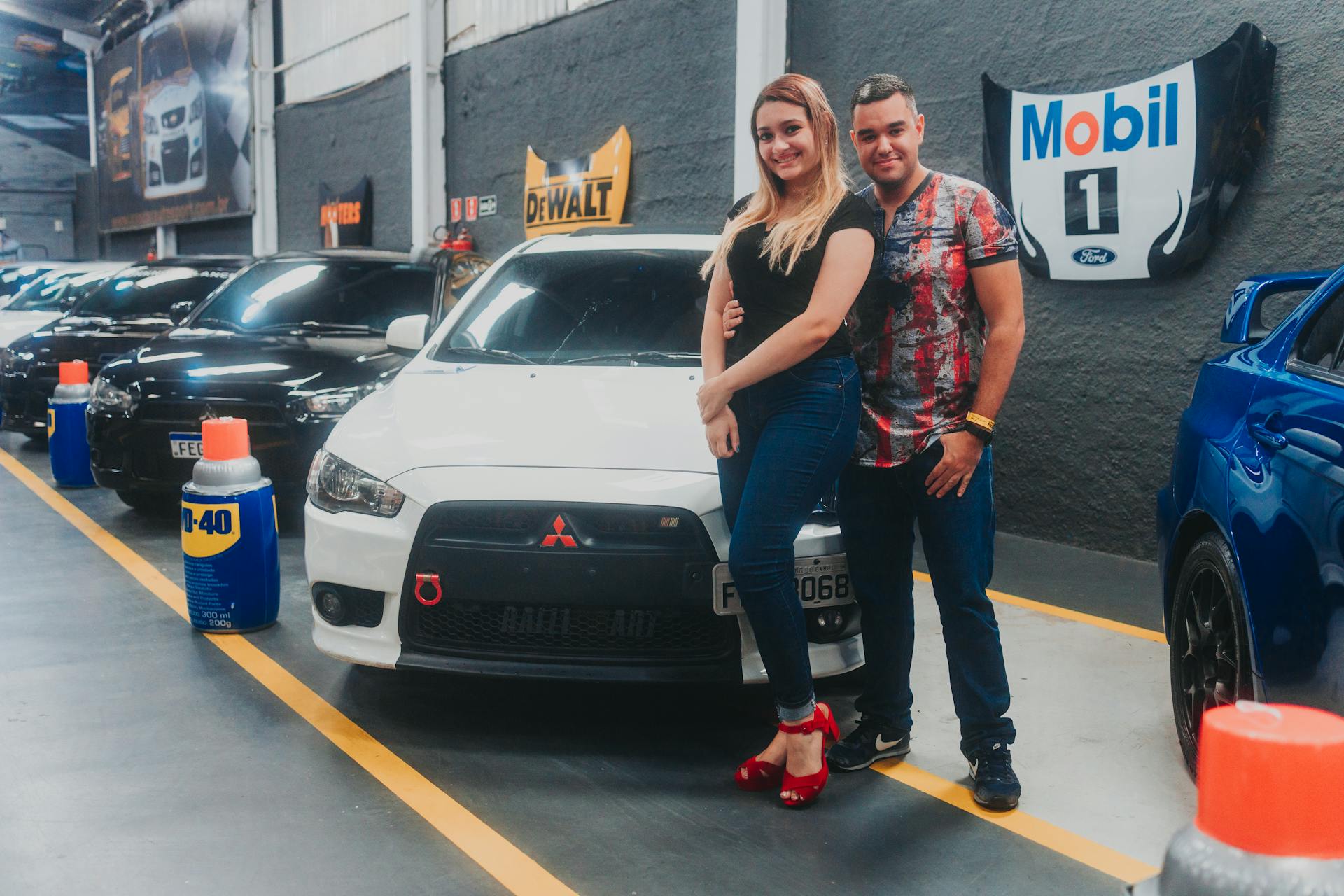 Smiling couple posing with white Mitsubishi Lancer at an indoor car show, surrounded by other vehicles.