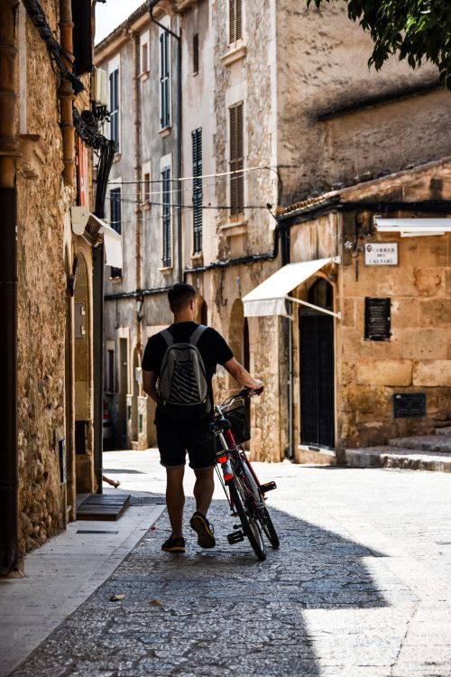 Free Back view of a man walking a bicycle in a quaint European alley, showcasing rustic architecture. Stock Photo